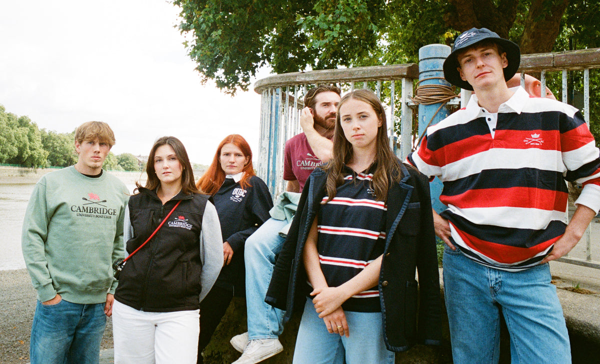 A group of six young adults posing outdoors by a riverbank next to a metal railing. They are wearing casual vintage-style Boat Race clothing, including a Cambridge Blue sweatshirt, a black vest, a striped polo shirt, and jeans.
