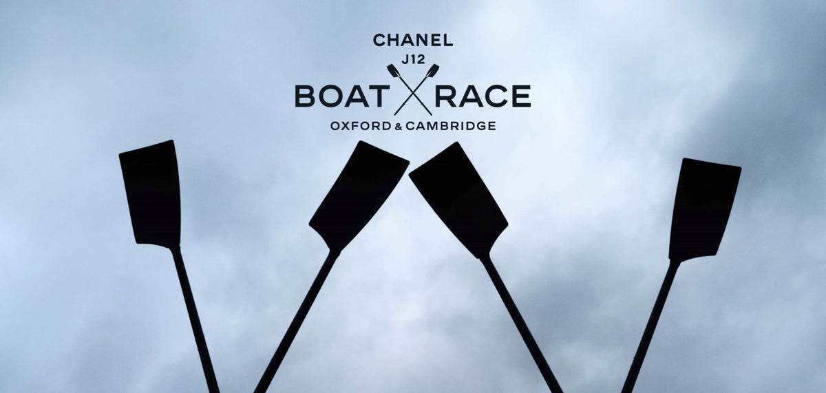 Chanel J12 Boat Race Oxford & Cambridge logo centered over four black oar silhouettes against a cloudy blue-gray sky.