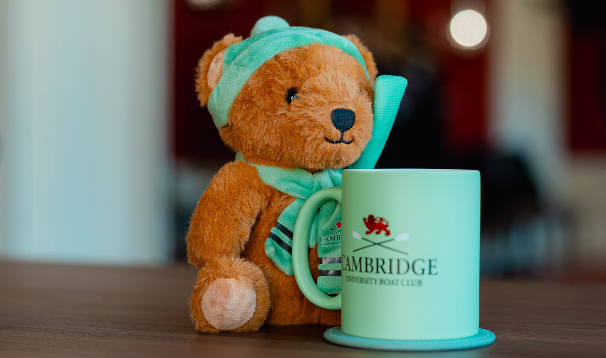 A small brown teddy bear wearing a Cambridge Blue hat and scarf sits next to a mint green ceramic mug with a matching coaster and spoon. The mug is printed with the Cambridge University Boat Club logo and text, and the background is a softly blurred indoor setting.