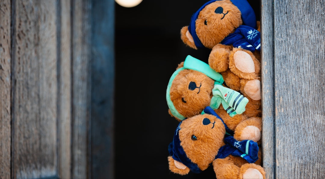Three small brown teddy bears wearing Blue and Cambridge Blue winter outfits and Rowing Oars peek out from behind a rustic wooden door.